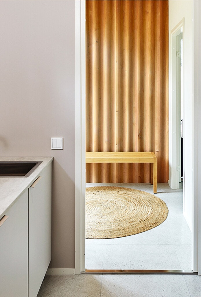 Greigen sävyiseksi sisustetun kodinhoitohuoneen ovensuusta näkyy saunan pukuhuoneen lämpimänsävyinen pystysuuntaan asennettu puinen sormipaneelitehosteseinä.
